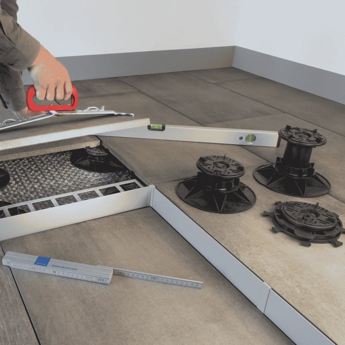 Ambientebild der 3 verschiedenen Stelzlagergrößen auf einer Terrasse. Der Handwerker benutzt eine Wasserwaage und einen Plattenheber zum Fliesenlegen.